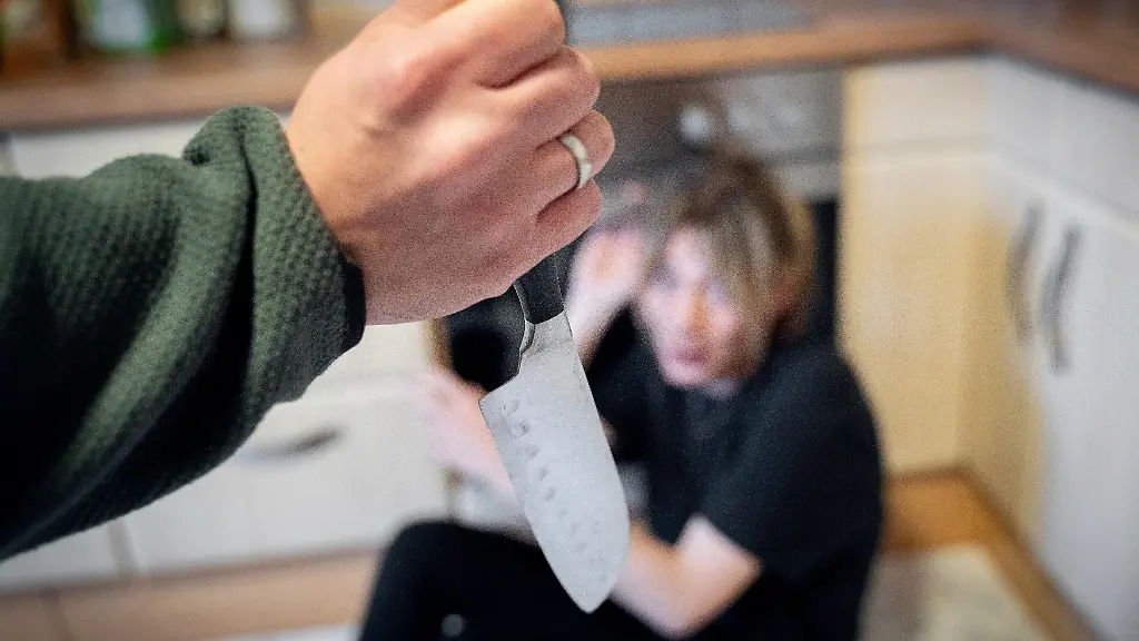 Ein-Mann-bedroht-eine-Frau-in-einer-Kueche-mit-einem-Messer-waehrend-sie-veraengstigt-am-Boden-sitzt-was-als-Symbolbild-fuer-haeusliche-Gewalt-Eskalation-in-Beziehungen-und-akute-Bedrohungssituationen-steht