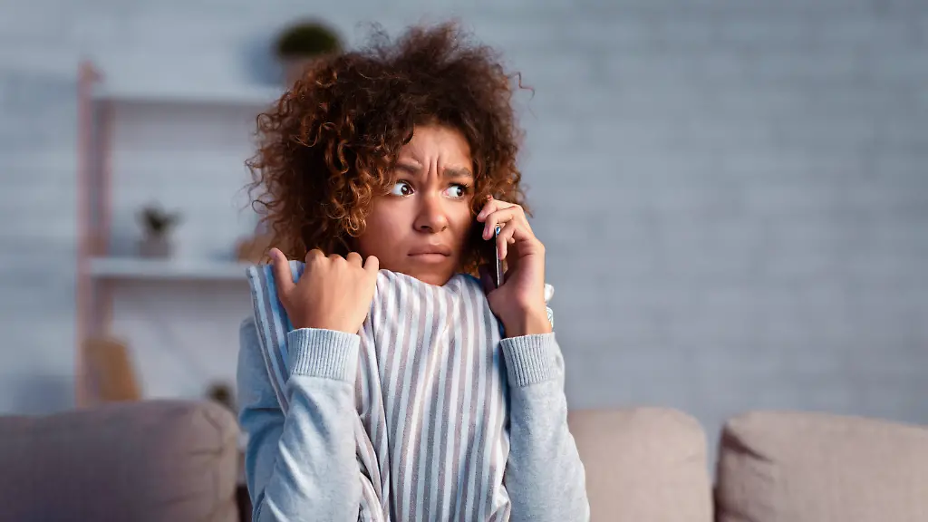 Scared-afro-woman-calling-phone-and-embracing-pillow-Scared-afro-woman-calling-phone-and-embracing-pillow-on-sofa-afraid-of-noise-near-house-free-space-model-released-61238388