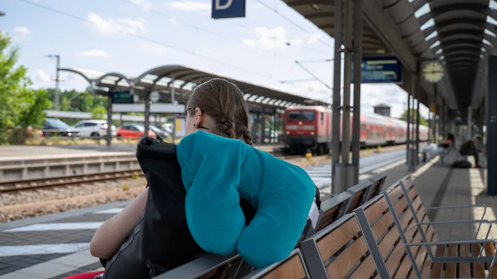 Symbolbild-Junge-Frau-mit-Rucksack-und-Nackenkissen-wartet-am-Bahnhof-29-06-2026-Eine-Reisende-sitzt-mit-Gepaeck-und-Nackenkissen-im-Bahnhofsbereich-und-wartet-auf-ihre-Zugverbindung