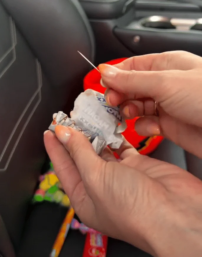 Eine Hand hält eine Nadel neben einer geöffneten Maoam-Verpackung; im Hintergrund steht ein Halloween-Kürbisbehälter.