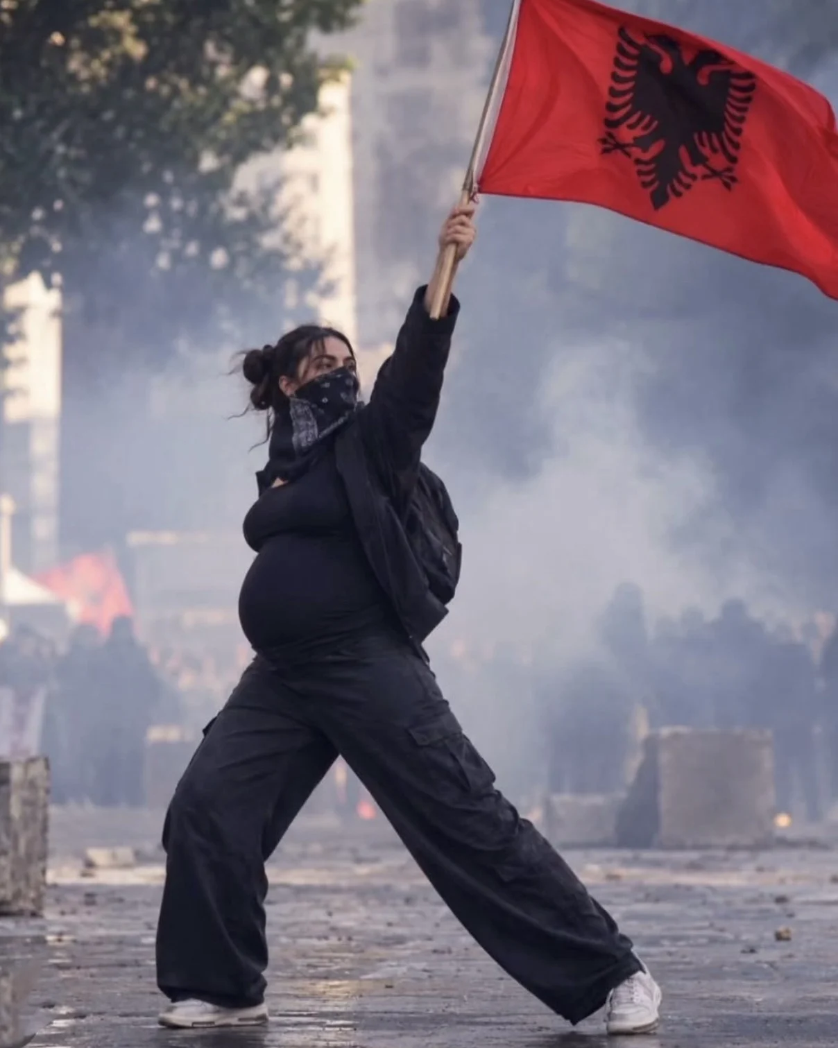 KI-generierte Darstellung einer schwangeren Frau mit albanischer Flagge vor einer rauchigen Straßenszene.